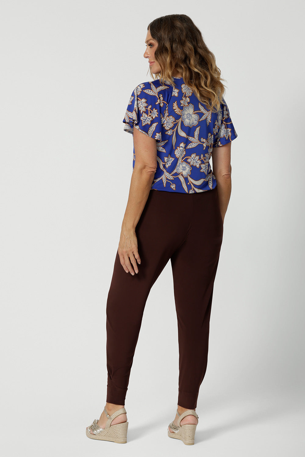 Back view of good off duty pants for tall, women, these tapered leg, long length pants by Australian and New Zealand women's  clothing brand, Leina & Fleur are comfortable stretchy pants in espresso brown jersey fabric. Worn with a flutter sleeve summer top in blue floral print jersey, these are easy care tall pants for summer style. Shop travel pants for plus size women and ladies tall pants online in sizes 8 to 24.