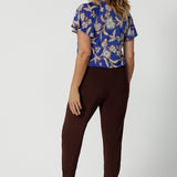 Back view of good off duty pants for tall, women, these tapered leg, long length pants by Australian and New Zealand women's  clothing brand, Leina & Fleur are comfortable stretchy pants in espresso brown jersey fabric. Worn with a flutter sleeve summer top in blue floral print jersey, these are easy care tall pants for summer style. Shop travel pants for plus size women and ladies tall pants online in sizes 8 to 24.