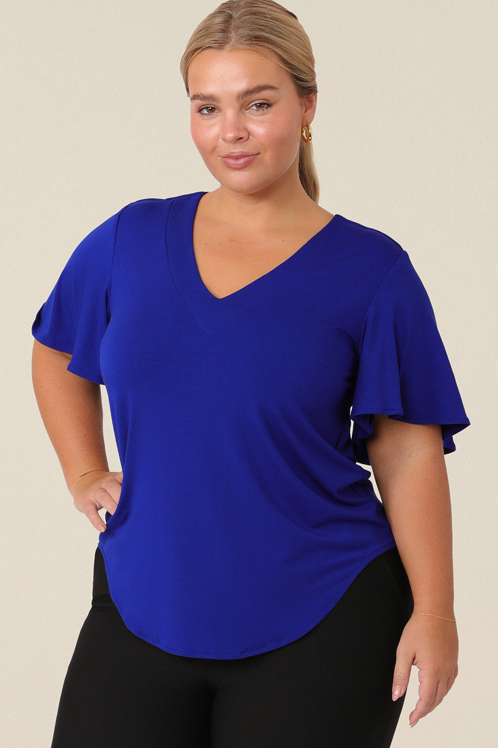 A plus size, size 14 woman wears a V-neck blue top with short flutter sleeves. Made from bamboo jersey, this is a natural fibre top that is lightweight, breathable and sustainable.