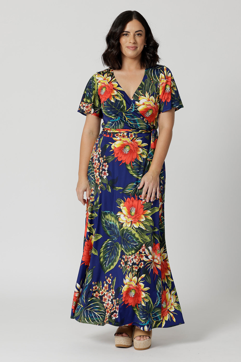 Maxi wrap dress worn by petite model in image. Made of easy care slinky jersey. Floral design depicts orange, yellow flowers, green and blue leaves on a deep blue base. Petite friendly dress come in sizes 8-24.