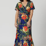 Maxi wrap dress worn by petite model in image. Made of easy care slinky jersey. Floral design depicts orange, yellow flowers, green and blue leaves on a deep blue base. Petite friendly dress come in sizes 8-24.