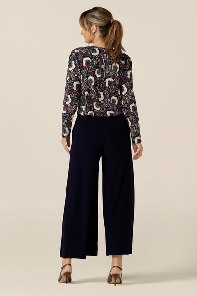 Back view of a size 10 woman wearing wide leg pull-on pants in navy stretch jersey with a paisley print, long sleeve cowl neck top. Comfortable pants for work and casual wear, these cropped trousers are made in Australia by women's clothing brand, Leina & Fleur.
