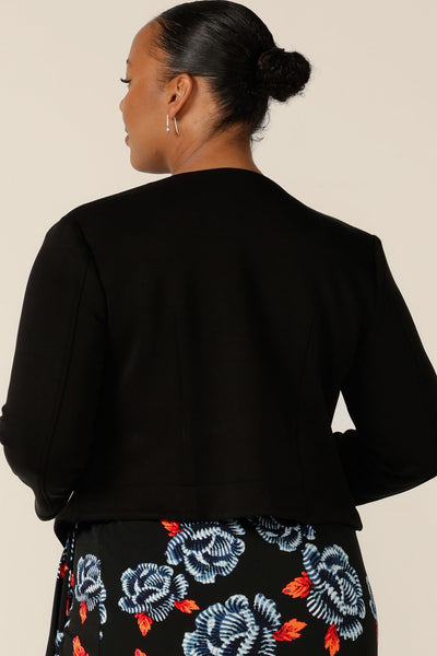 Back view of a classic work jacket, the Yuri Jacket in Black is a collarless, open-front, soft tailored jacket. Shown here in a size 18, the jacket is worn with a floral print, jersey wrap dress. Both are made in Australia by Australian and New Zealand women's clothing label, L&F. 