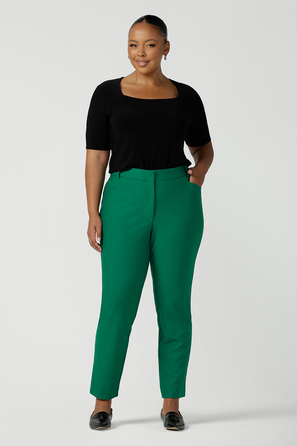 A curvy woman wears a plus size, short sleeve black top with square neckline. A slim fit top in classic black, it is worn with emerald green tapered, cigarette pants. This tailored jersey top wears well with workwear separates for an office look and also as a smart casual top. Shop tops in petite, mid size and plus size online at Australian women's clothing brand, Leina & Fleur.