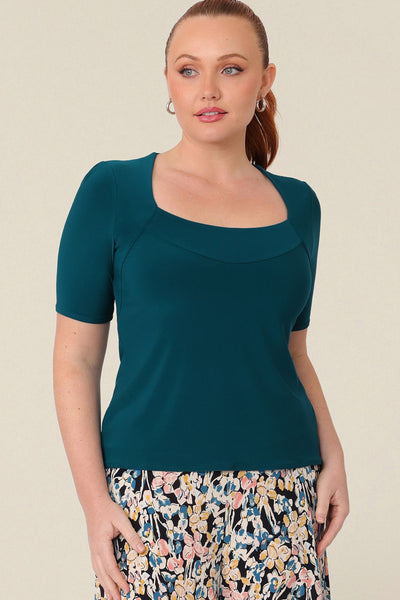 A size 12 woman wears a short-sleeve jersey top in dark teal. The top has a squared scoop neckline, a structured bust and short sleeves.  Wear for work or casual and shop in sizes 8 to size 24.