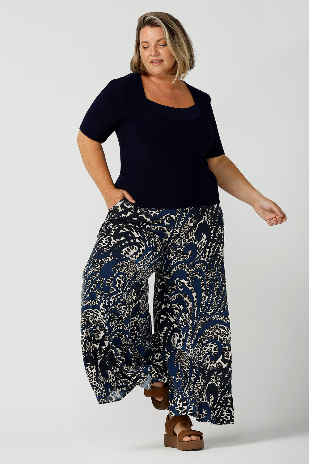 A petite height size, size 16 woman wears a casual jersey top in navy blue, styled with animal print, wide leg pants. This Australian-made women's top has short fitted sleeves, a square neckline and and high-low - perfect for weekend casual and travel capsule wardrobes.