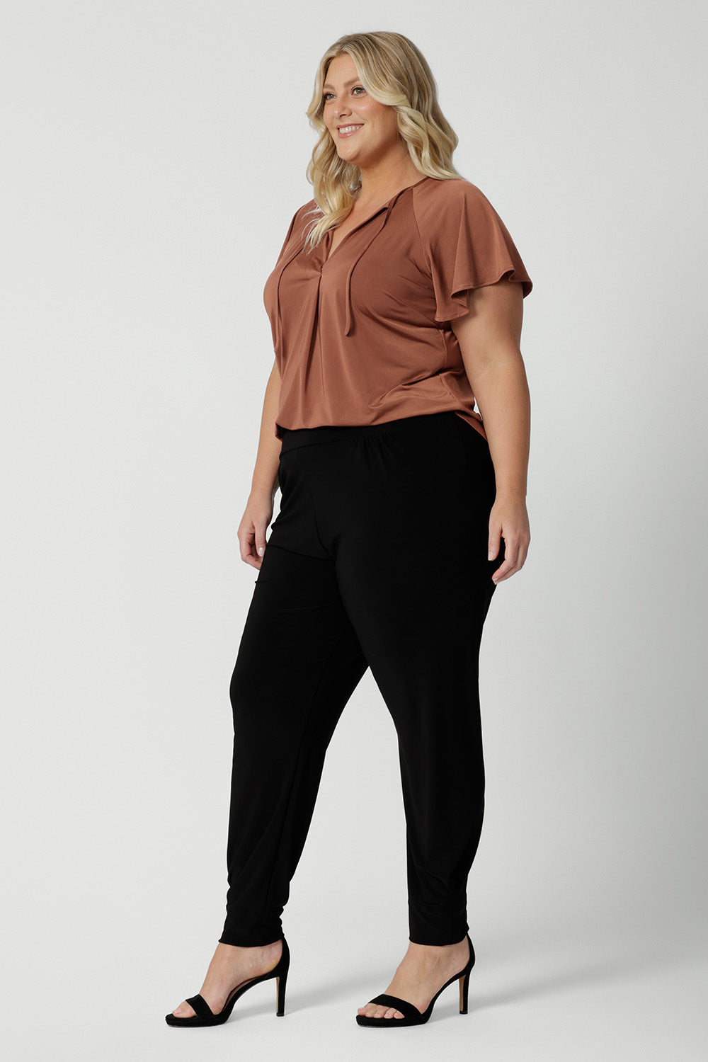 A good summer top for over plus size women, a size 18 woman wears a flutter sleeve top in clay slinky jersey fabric. An easy-care top for travel and cruise wear, this made-in-Australia V neck top is worn with tapered leg black travel pants.  By Australian and New Zealand women's clothes label, Leina and Fleur shop petite to plus size tops for women  online.