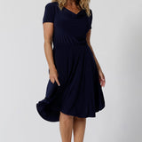 Navy dresses for over 40s women don't get much beter than this! A size 12, over 40 woman wears knee length summer dress with asymmetric hemlineand cowl neck. A short sleeve dress in navy jersey fabric, this is an easy-care dress and makes a good jersey dress for travel and cruise wear.  Made in Australia by Australian and New Zealand women's clothes brand, Leina & Fleur, shop their petite to plus size dresses online. 