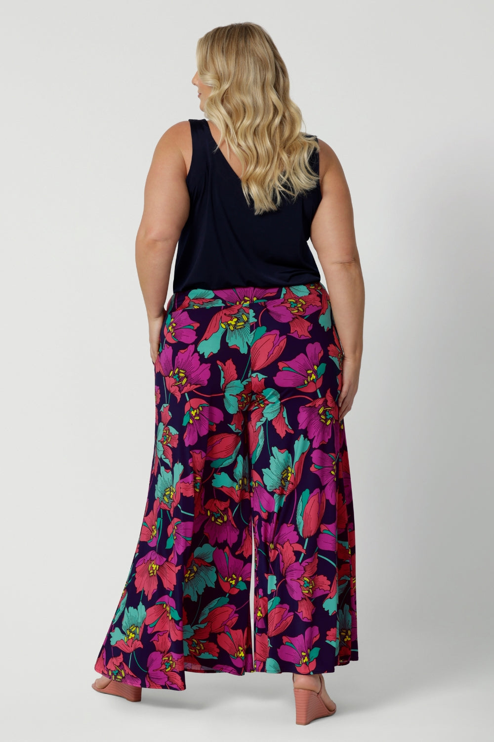 An image of the back of a curvy size 18 woman wearing the Caspian Palazzo pant in a bright floral print. In soft and lightweight jersey this pant features side pockets and a flared leg opening. Made in sizes 8 to 24 by Australian fashion brand Leina & Fleur.