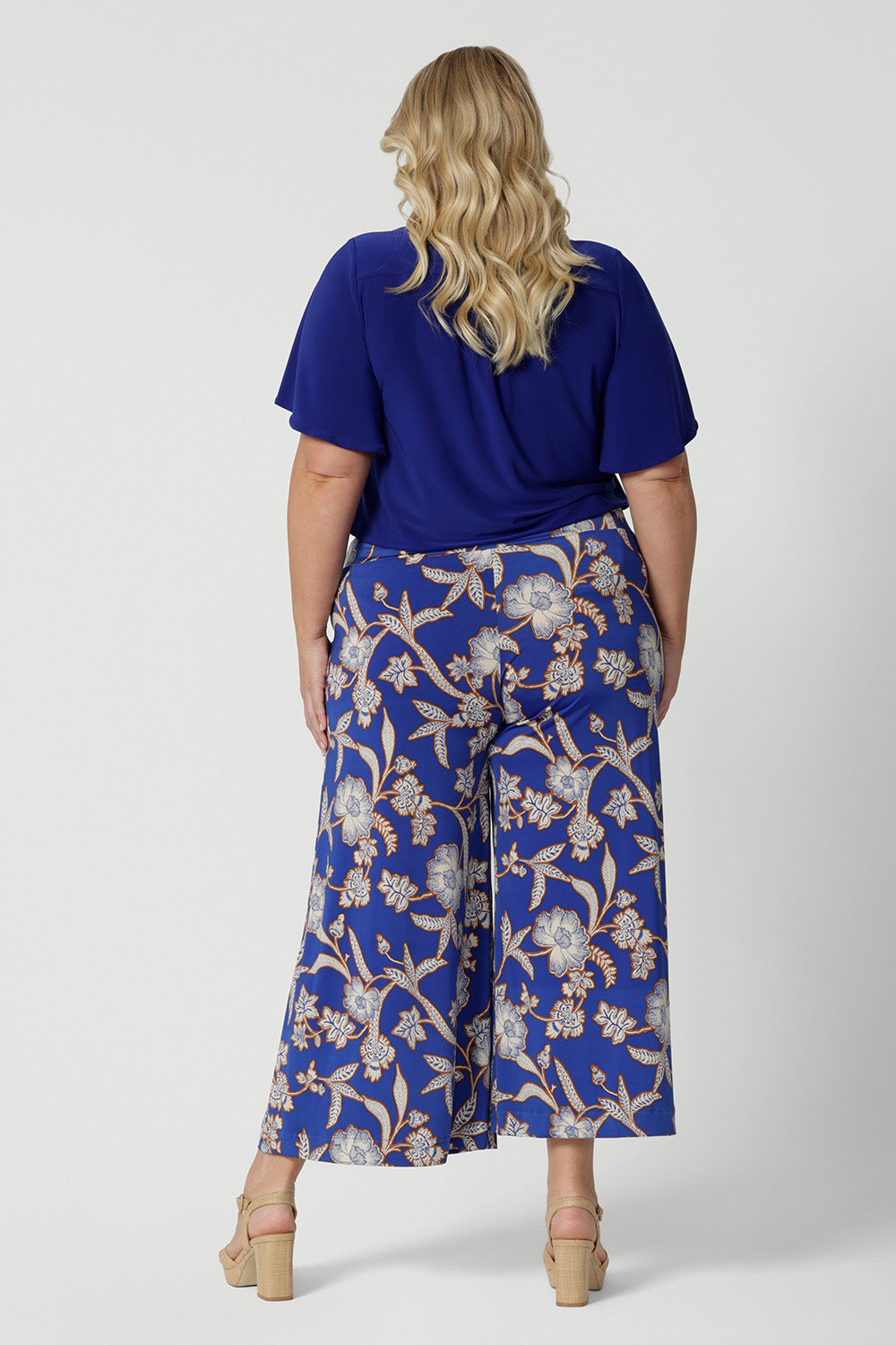 Wide leg pants for summer don't come much better than this! Worn by a size 18, curvy woman, this is a back view of plus size wide leg pants are in easy care jersey with a blue, caramel and white floral print. Made in Australia by Australian and New Zealand women's clothes brand, Leina and Fleur. Shop wide leg pants in petite to plus sizes.