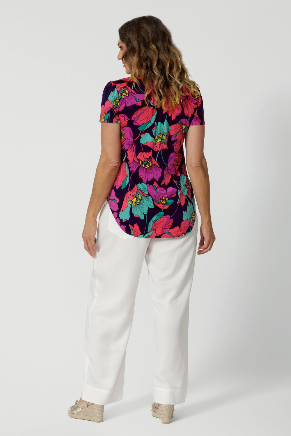 The back of a curvy size 10 woman over 40 wears a top with a v-neckline and short sleeves. It has a bold floral print. She has styled the top with white resort pants. The clothes are handmade in Australia by Australian fashion brand Leina & Fleur in sizes 8 to 24. 