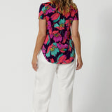 The back of a curvy size 10 woman over 40 wears a top with a v-neckline and short sleeves. It has a bold floral print. She has styled the top with white resort pants. The clothes are handmade in Australia by Australian fashion brand Leina & Fleur in sizes 8 to 24. 