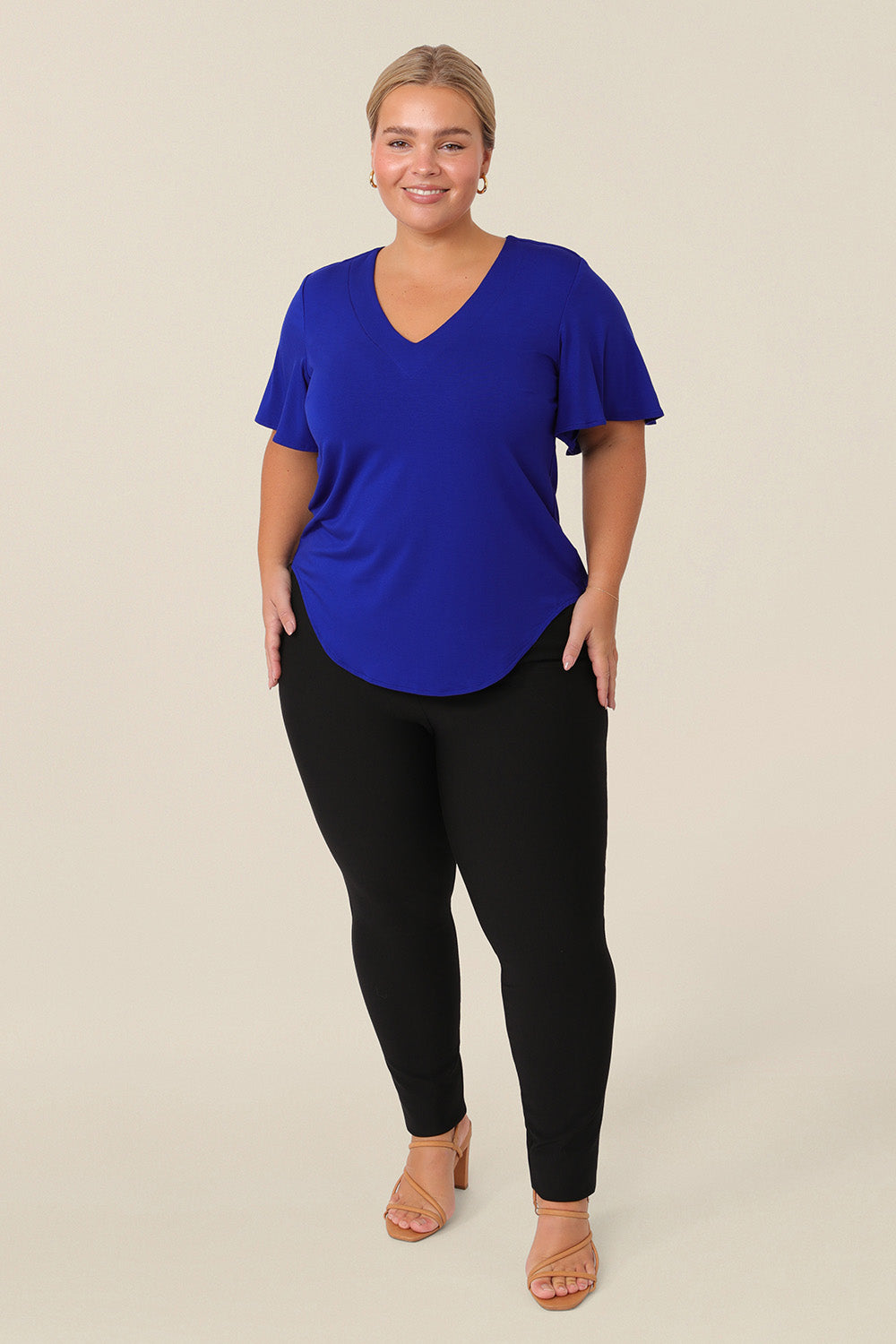 A petite height, size 14 woman wears a V-neck blue top with short flutter sleeves. Made from bamboo jersey, this is a natural fibre top that is lightweight, breathable and sustainable. The blue bamboo jersey top is worn over slim-leg black jersey trousers for a smart-casual look.