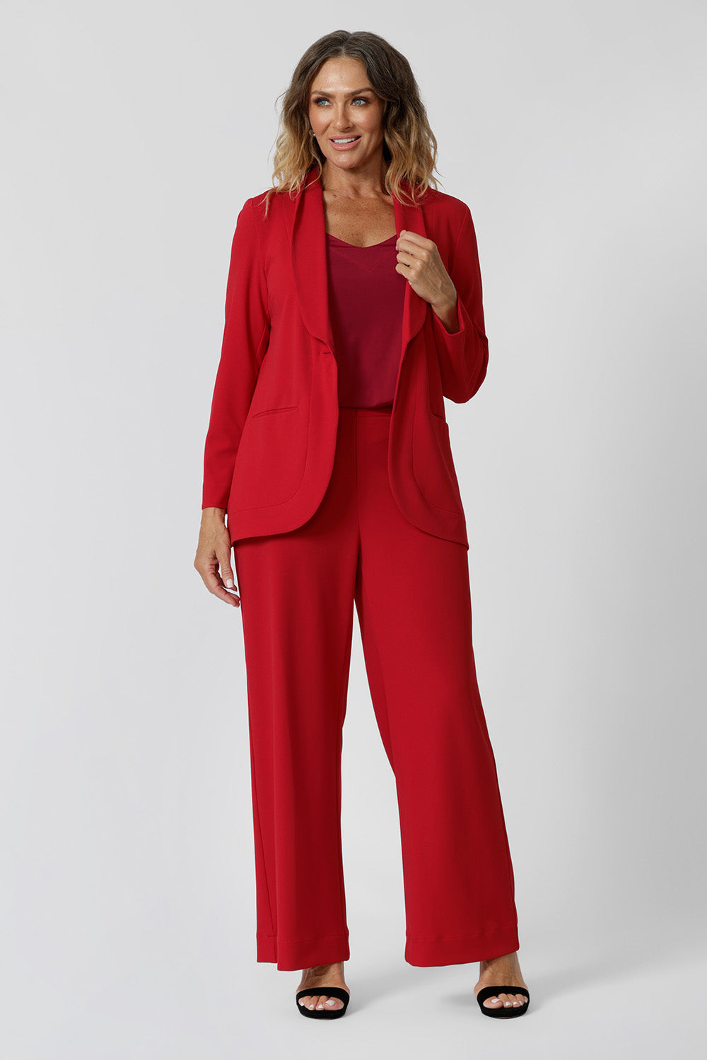 A size 12 woman wears the Merit Blazer in Flame, a textured scuba crepe blazer with button front. This red work blazer is worn with red workwear pants and red cami top. Curved hemline and front pockets. Made in Australia for women size 8 - 24.