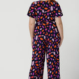 Back view of a size 12 woman wears the Blanca Jumpsuit in Palette. A wrap jumpsuit design with a flutter sleeve, navy base colour and vibrant fuchsia, red, purple brush strokes throughout the print. Cropped leg length and petite to plus size friendly. Made in Australia for women size 8 - 24.
