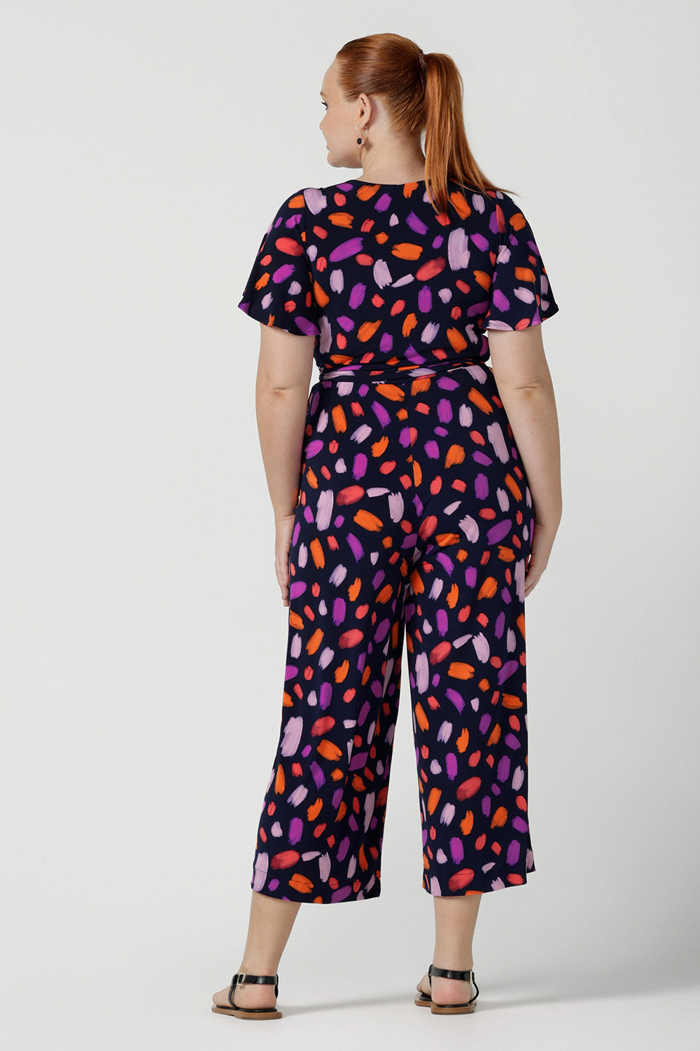 Back view of a size 12 woman wears the Blanca Jumpsuit in Palette. A wrap jumpsuit design with a flutter sleeve, navy base colour and vibrant fuchsia, red, purple brush strokes throughout the print. Cropped leg length and petite to plus size friendly. Made in Australia for women size 8 - 24.