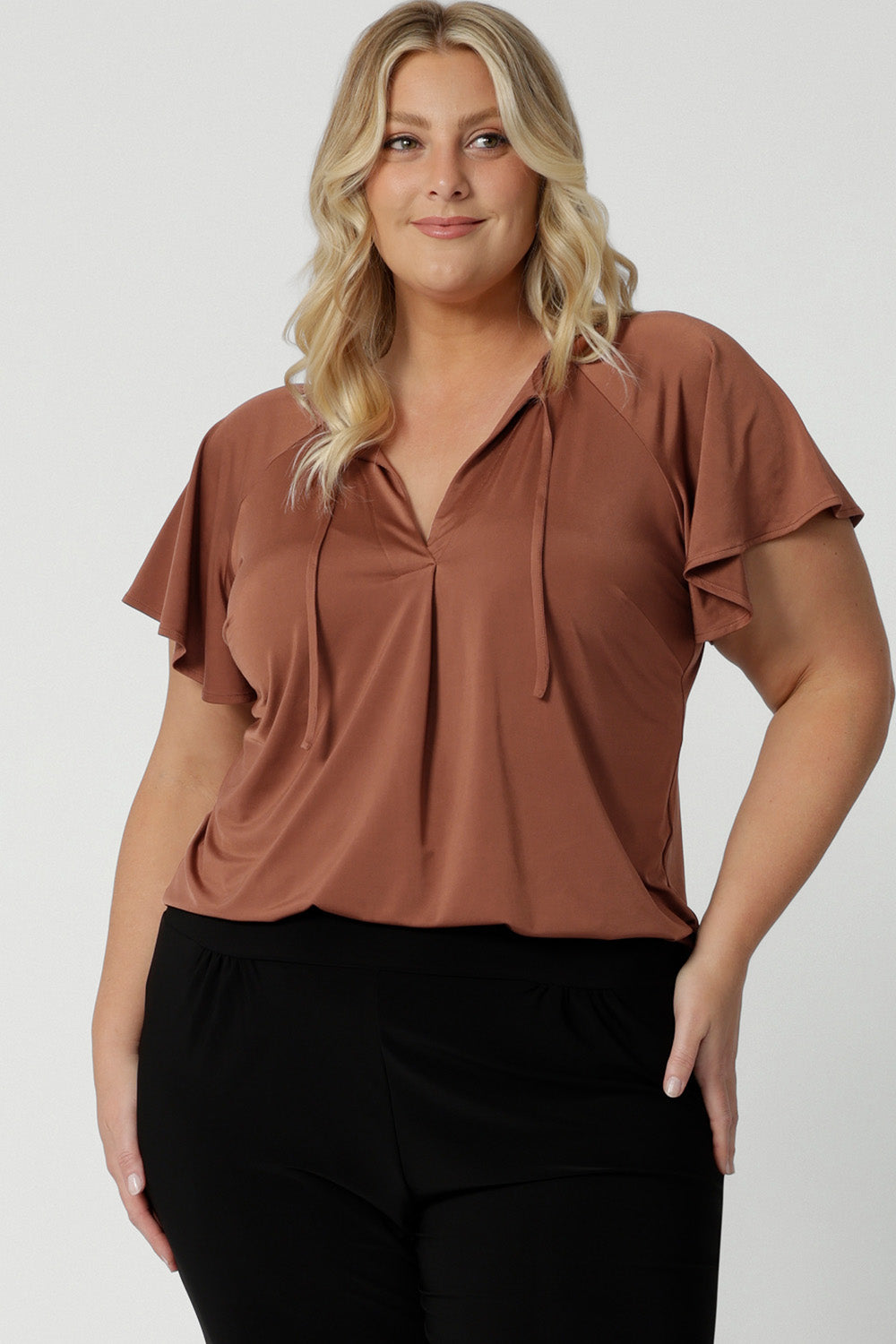 A good summer top for curvy women, a size 18 woman wears a plus size flutter sleeve top in clay slinky jersey fabric. An easy-care top for travel and cruise wear, this made-in-Australia V neck top is worn with tapered leg black travel pants.  By Australian and New Zealand women's clothing brand, Leina and Fleur shop petite to plus size tops for women  online.