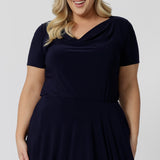 Summer dresses for curvy women don't get much beter than this! A size 18, curve woman wears plus size, knee length summer dress with asymmetric hemlineand cowl neck. A short sleeve dress in navy jersey fabric, this is an easy-care dress and makes a good jersey dress for travel and cruise wear.  Made in Australia by Australian and New Zealand women's clothes brand, Leina & Fleur, shop their petite to plus size dresses online. 