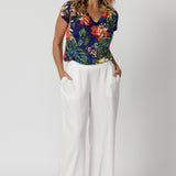 Full body image of 40 year old woman wearing floral top with a cowl neck detail paired with white pants. This easy care top is made from slinky jersey fabric with a colourful combination of orange, yellow and white flowers on a deep blue base.  Designed in Australia for Australian and New Zealand women. Leina & Fleur is a woman's clothing brand that stock sizes from 8 to 24. 