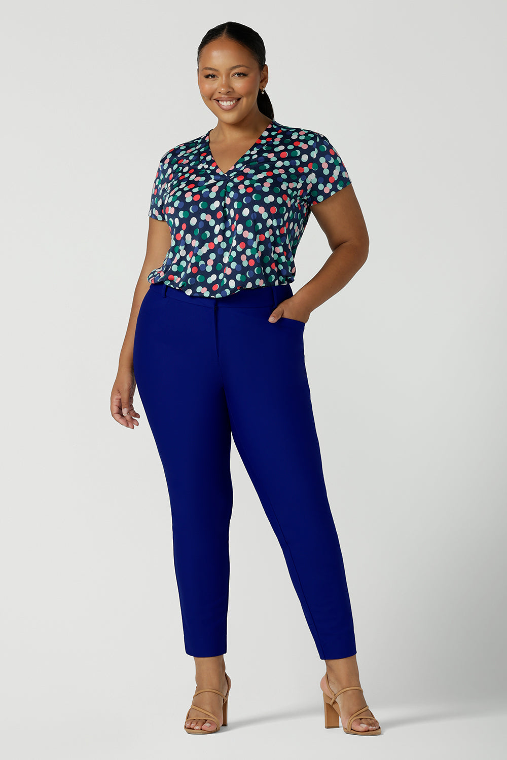A plus size, size 18 woman wears a short sleeve, V-neck top in dot print slinky jersey fabric. Worn with cobalt blue tailored trousers, this women's top is a good top for plus size women's workwear with personality! Made in Australia and available to shop online in sizes 8 to 24.