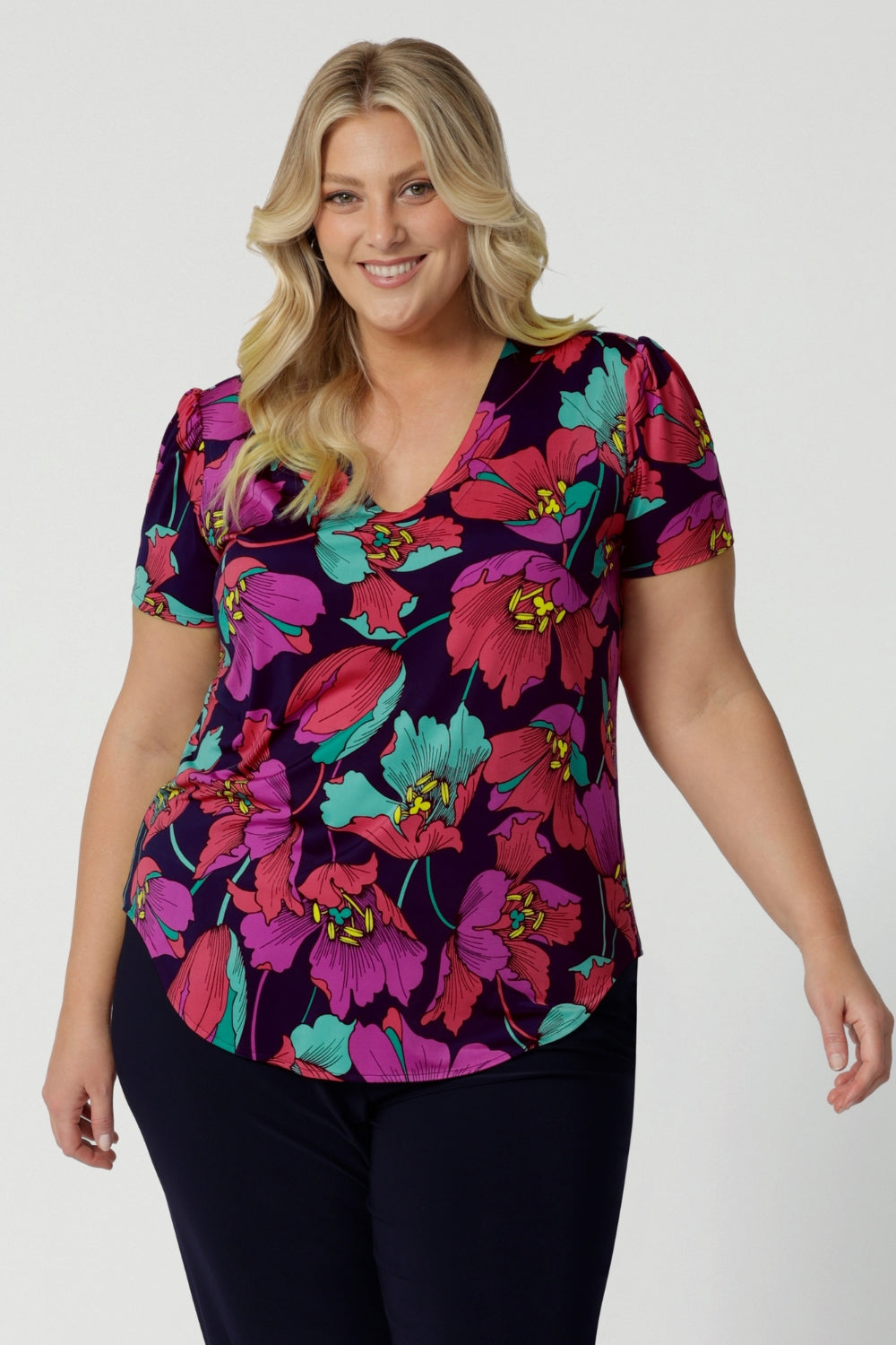 A curvy size 18 woman over 35 wears a top with a v-neckline and short sleeves. It has a bold floral print. The top is handmade in Australia by Australian fashion brand Leina & Fleur in sizes 8 to 24. 