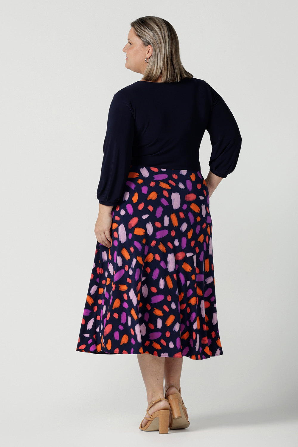 Back view of a size 18 curvy woman wears the Nerida dress. Functioning fit and flare wrap dress with a navy bodice and printed skirt. 3/4 Sleeves and functioning pockets. Made in Australia for women. Size 8 - 24. Styled back with a brown sling back heel.