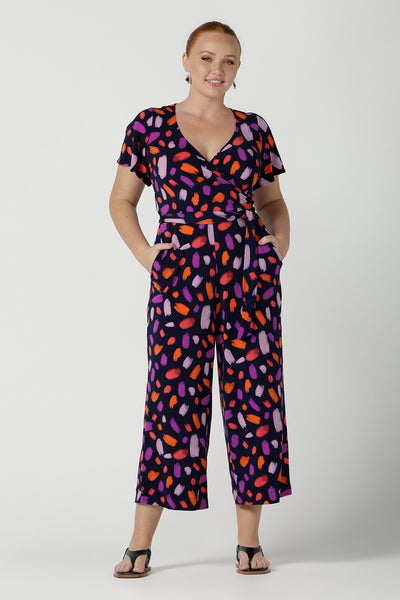 Size 12 woman wears the Blanca Jumpsuit in Palette. A wrap jumpsuit design with a flutter sleeve, navy base colour and vibrant fuchsia, red, purple brush strokes throughout the print. Cropped leg length and petite to plus size friendly. Made in Australia for women size 8 - 24.
