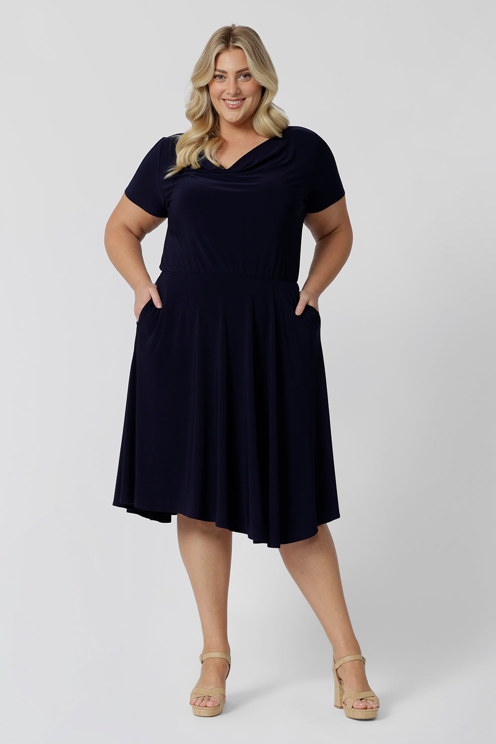 Summer dresses for curvy women don't get much beter than this! A size 18, curve woman stands with her hands in the pockets of a plus size, knee length summer dress with asymmetric hemlineand cowl neck. A short sleeve dress in navy jersey fabric, this is an easy-care dress and makes a good jersey dress for travel and cruise wear.  Made in Australia by Australian and New Zealand women's clothes brand, Leina & Fleur, shop their petite to plus size dresses online. 