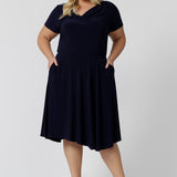 Summer dresses for curvy women don't get much beter than this! A size 18, curve woman stands with her hands in the pockets of a plus size, knee length summer dress with asymmetric hemlineand cowl neck. A short sleeve dress in navy jersey fabric, this is an easy-care dress and makes a good jersey dress for travel and cruise wear.  Made in Australia by Australian and New Zealand women's clothes brand, Leina & Fleur, shop their petite to plus size dresses online. 