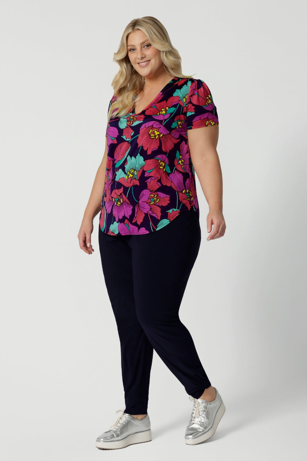 A curvy size 18 woman over 35 wears a top with a v-neckline and short sleeves. It has a bold floral print. She has styled the top with navy stretchy travel pants. The clothes are handmade in Australia by Australian fashion brand Leina & Fleur in sizes 8 to 24. 