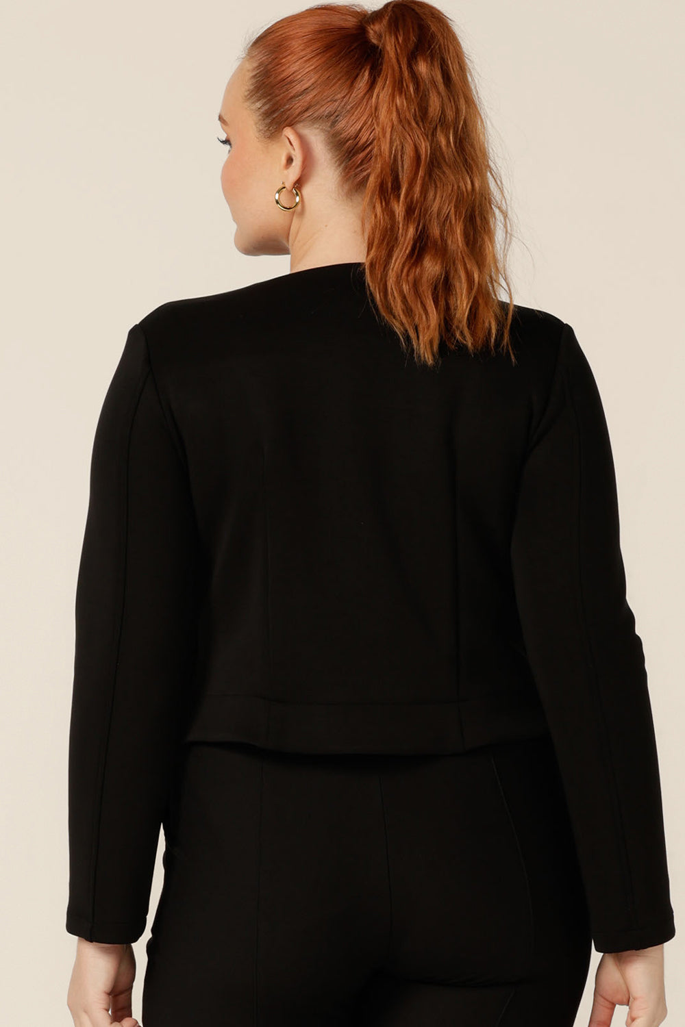 Back view of a classic workwear jacket by Australian and New Zealand women's clothing label, L&F. The Yuri Jacket in Black is a collarless, open-front, soft tailored jacket. Shown here in a size 12, the jacket is worn with black tailored pants to create a work suit look, and a V-neck top in orange. 