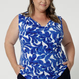 Plus sized woman wears sleeveless top, crafted from a lightweight, dry-touch jersey fabric, making it perfect for warm weather and upcoming summer events. The vibrant cobalt blue colour is complemented by delicate white leaf detailing, adding a touch of nature-inspired elegance. Easy care top is made all shapes and sizes in mind by clothing brand Leina & Fleur in sizes 8-24.