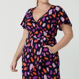 Close up of a size 12 woman wears the Blanca Jumpsuit in Palette. A wrap jumpsuit design with a flutter sleeve, navy base colour and vibrant fuchsia, red, purple brush strokes throughout the print. Cropped leg length and petite to plus size friendly. Made in Australia for women size 8 - 24.