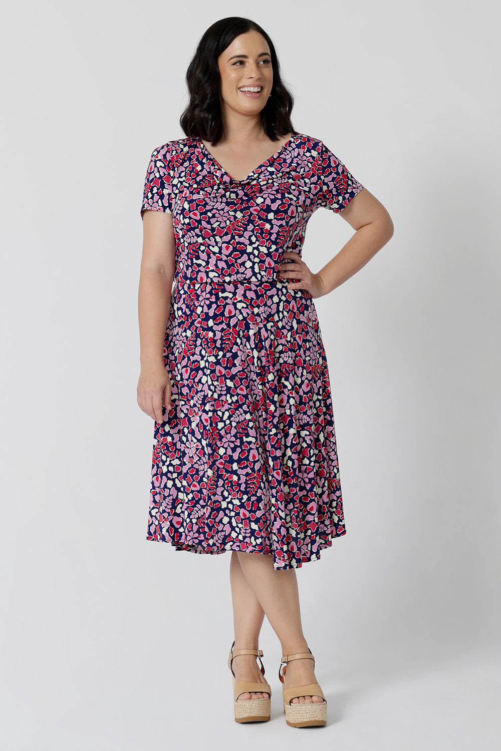 Summer dresses for petite women don't get much beter than this! A size petite size 10 woman wears knee length summer dress with asymmetric hemline and cowl neck. A short sleeve dress in pretty red, pink, white and navy print this is a good jersey dress for travel and cruise wear.  Made in Australia by Australian and New Zealand women's clothes brand, Leina & Fleur, shop their petite to plus size dresses online. 