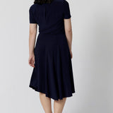 Back view oa a navy dress for petite women. A petite size 10 woman wears knee length summer dress with asymmetric hemlineand cowl neck. A short sleeve dress in navy jersey fabric, this is an easy-care dress and makes a good jersey dress for travel and cruise wear.  Made in Australia by Australian and New Zealand women's clothes brand, Leina & Fleur, shop their petite to plus size dresses online. 