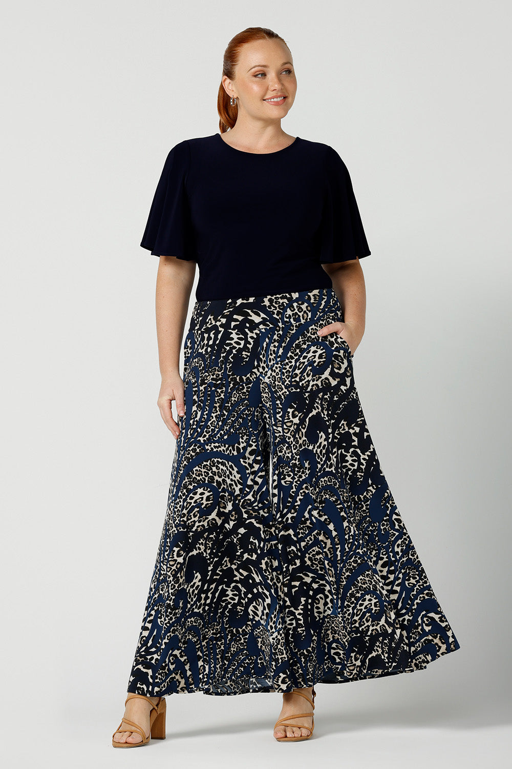 A curvy size, size 12 woman wears printed wide leg pants with pockets with a navy round neck top. These pull-on, easy care pants are comfortable for your everyday workwear capsule wardrobe. Shop these Australian-made wide leg trousers online in sizes 8 to 24, petite to plus sizes.