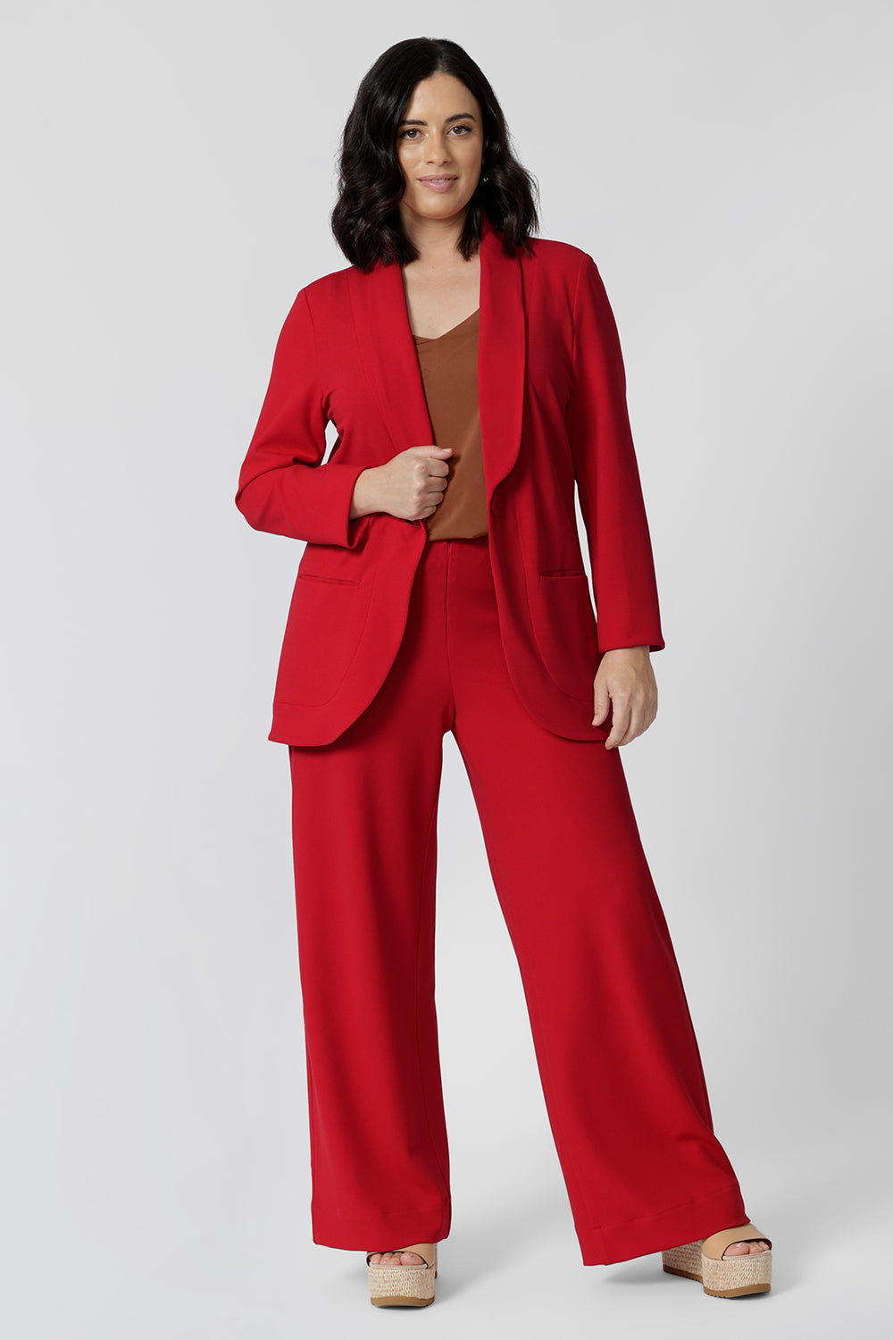 A petite height woman wears the Merit Blazer in Flame, a textured scuba crepe plus size blazer with button front. This red work blazer is worn with red workwear pants and navy work top. Curved hemline and front pockets. This blazer is Made in Australia for petite women in sizes 8 - 24.