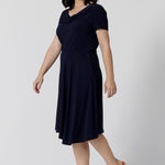 Navy dresses for petite women don't get much beter than this! A petite size 10 woman wears knee length summer dress with asymmetric hemlineand cowl neck. A short sleeve dress in navy jersey fabric, this is an easy-care dress and makes a good jersey dress for travel and cruise wear.  Made in Australia by Australian and New Zealand women's clothes brand, Leina & Fleur, shop their petite to plus size dresses online. 