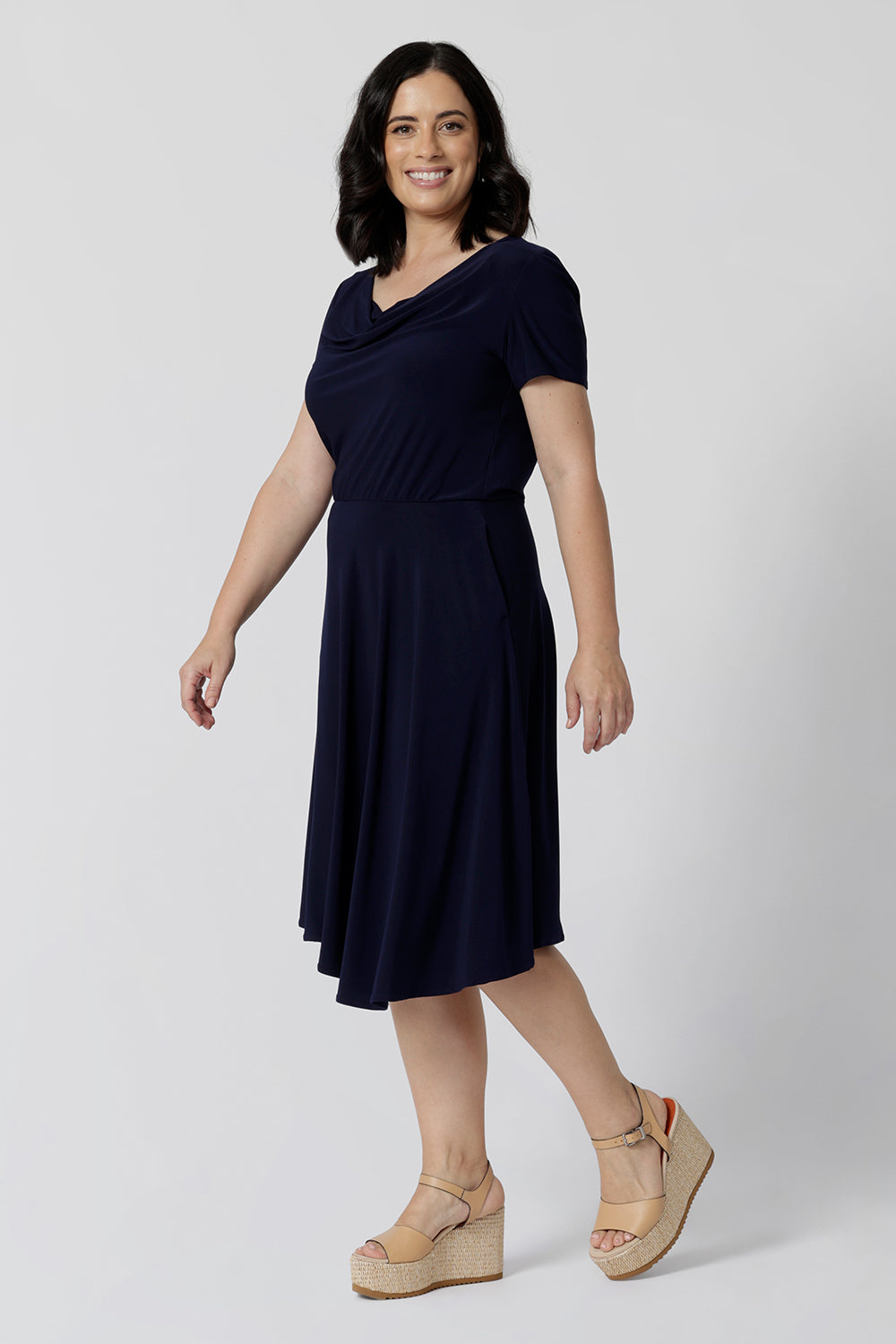 Navy dresses for petite women don't get much beter than this! A petite size 10 woman wears knee length summer dress with asymmetric hemlineand cowl neck. A short sleeve dress in navy jersey fabric, this is an easy-care dress and makes a good jersey dress for travel and cruise wear.  Made in Australia by Australian and New Zealand women's clothes brand, Leina & Fleur, shop their petite to plus size dresses online. 