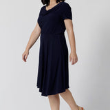 Navy dresses for petite women don't get much beter than this! A petite size 10 woman wears knee length summer dress with asymmetric hemlineand cowl neck. A short sleeve dress in navy jersey fabric, this is an easy-care dress and makes a good jersey dress for travel and cruise wear.  Made in Australia by Australian and New Zealand women's clothes brand, Leina & Fleur, shop their petite to plus size dresses online. 