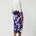 Back view of a size 10 woman wearing the Aaron Midi Skirt in Jacaranda. A printed Midi length skirt in purple florals. Made in Australia for women size 8-24. Styled back with pink pumps and a purple Jacaranda jacket.