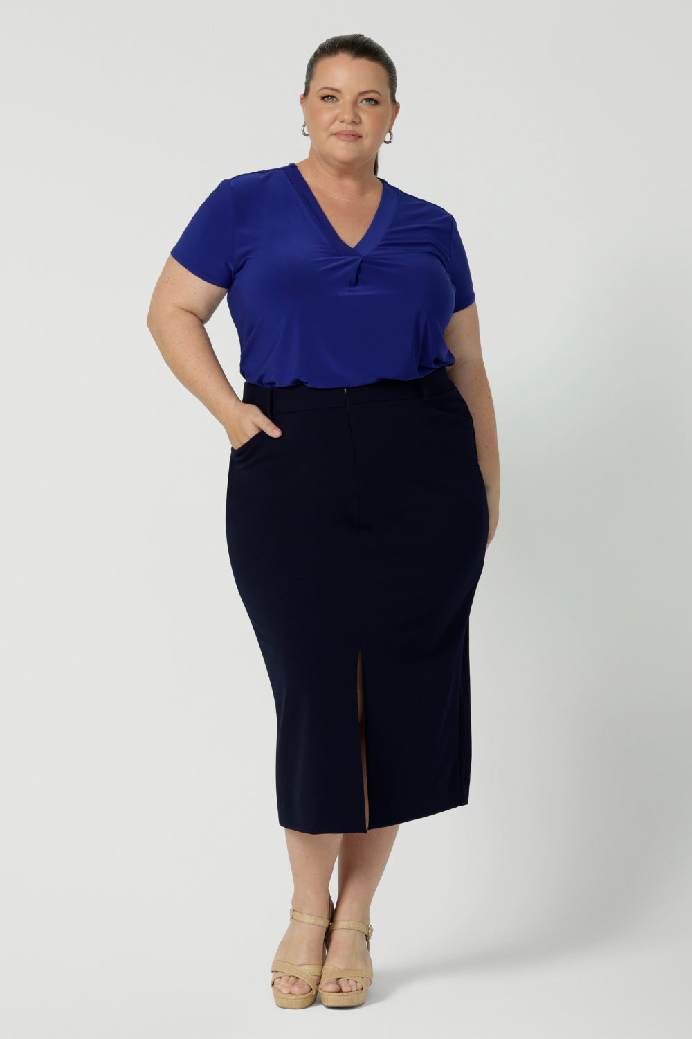 Full body front view of 40 year old plus sized woman wearing a midi length work skirt. This workwear skirt is navy in colour and made of good quality scuba crepe fabric. This skirt has an above the knee slit for comfort and mobility. Perfect skirt for an office or corporate setting. 3/4 length skirt is Australia made by New Zealand and Australian women. Leina & Fleur is a clothing company for woman that stock sizes from 8 to 24. Shop this work skirt in plus sizes and petite sizes. 