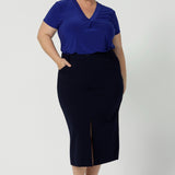 Full body front view of 40 year old plus sized woman wearing a midi length work skirt. This workwear skirt is navy in colour and made of good quality scuba crepe fabric. This skirt has an above the knee slit for comfort and mobility. Perfect skirt for an office or corporate setting. 3/4 length skirt is Australia made by New Zealand and Australian women. Leina & Fleur is a clothing company for woman that stock sizes from 8 to 24. Shop this work skirt in plus sizes and petite sizes. 