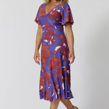 An image of a size 12, over 40 woman wears an empire line midi dress with flutter sleeves and twist front bodice. A good dress for wedding guest outfits, bold print makes it a pretty summer dress. Made in Australia by Australian and New Zealand women's clothes brand, Leina & Fleur, shop their petite to plus size dresses online. 