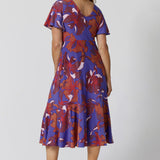 An image of size 12, over 40 woman wears an empire line midi dress with flutter sleeves and twist front bodice. She is facing backwards to show the v-neckline at the back of the dress. A good dress for wedding guest outfits, bold print makes it a pretty summer dress. Made in Australia by Australian and New Zealand women's clothes brand, Leina & Fleur, shop their petite to plus size dresses online. 