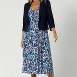 Size 12 woman wears flattering and functional wrap dress by Leina & Fleur. Featuring a double-layered back bodice for structure and sophistication, while the extended waist ties allow for a customisable fit. Model pairs dress with cropped navy jacket to create an elevated workwear outfit, tying together the navy with the blue hues in the midi dress.