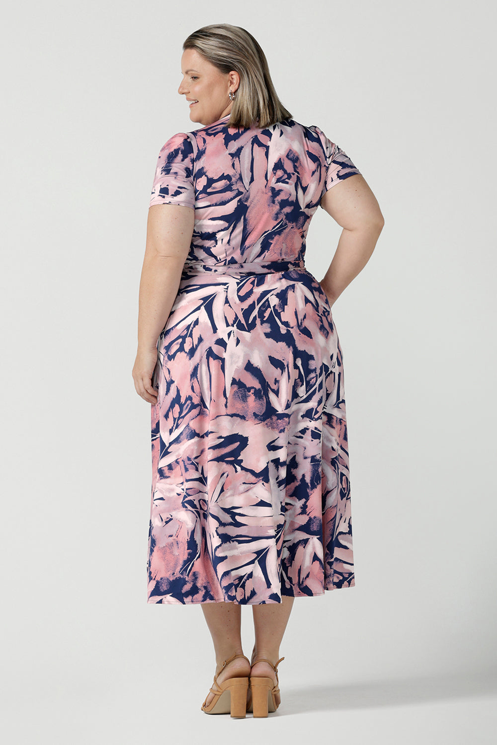 Back view of a size 18 Curvy woman wears the Alexis dress in the Cantata print. A functioning wrap dress with tie side and short sleeve and midi length. Navy base with an abstract brush strokes leafy print. Made in Australia for women size inclusive 8 - 24.