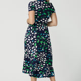 Back view of a size 12 woman wearing a jersey wrap dress in navy blue, green and white print. An easy-care work dress for everyday office and corporate wear, in sizes 8 to 24 this is a good plus size dress. Made in Australia by Australian and New Zealand womens clothing label, Leina & Fleur, this wrap dress is part of a collection of dresses for plus size to petite height women.  