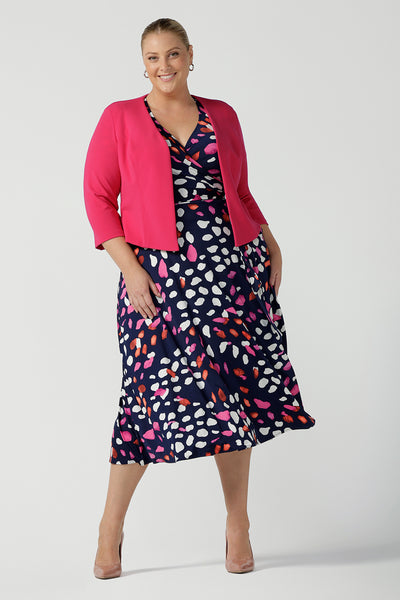 A curvy, size 18 woman wearing an abstract jersey print, wrap dress with a pink jacket. A great dress for summer casual wear, or for travel. Shop made in Australia dresses in petite to plus sizes online at Australian fashion brand, Leina & Fleur.