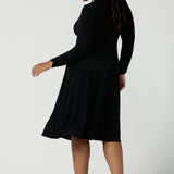 Back view of a size 16 woman wears a Alyssa Dress in jersey. V-neckline style great for work to weekend wear. Corporate dress for women. Travel friendly with an empire line style. Midi dress and styled back with black strappy heels. Made in Australia for women size 8 - 24.